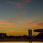 Vista panorâmica da Esplanada dos Ministérios em Brasília ao entardecer. O céu ocupa grande parte da imagem, com tons de azul, laranja e dourado refletidos pelas nuvens. Na parte inferior, aparecem vias urbanas com postes de iluminação, veículos e edifícios institucionais em silhueta. Ao centro, destacam-se duas torres retangulares do Congresso Nacional ao fundo. À direita, é visível parte de uma estrutura arquitetônica moderna de curvas marcantes, com um espelho d’água em primeiro plano refletindo as cores do pôr do sol. No canto superior esquerdo há um logotipo branco dentro de um quadrado com cantos arredondados. A cena transmite um aspecto institucional e representativo da capital federal brasileira.
